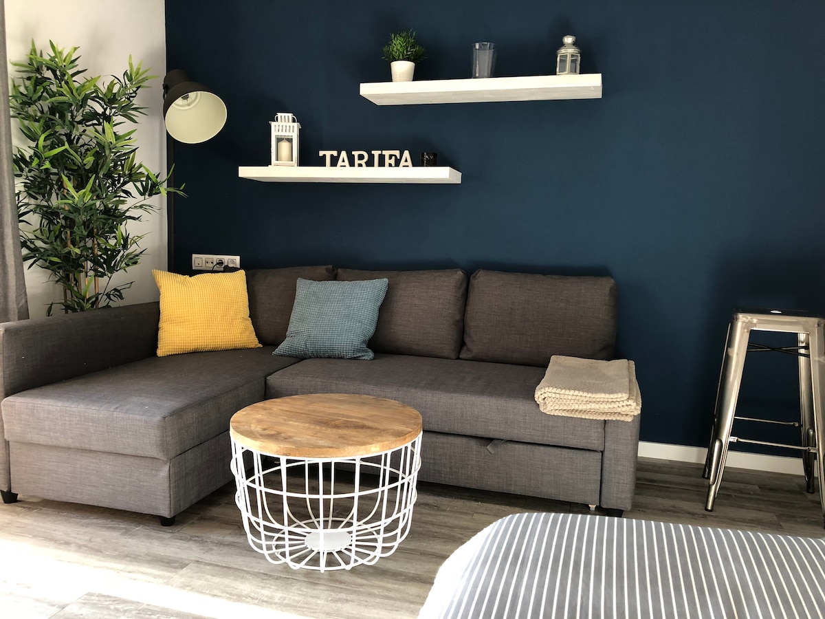 A modern living area features a sectional gray sofa adorned with colorful cushions in yellow and blue. Light wood accents are present in the round coffee table with a wire base. The blue wall is complemented by floating shelves showcasing decorative items, enhancing the space's cozy aesthetic.