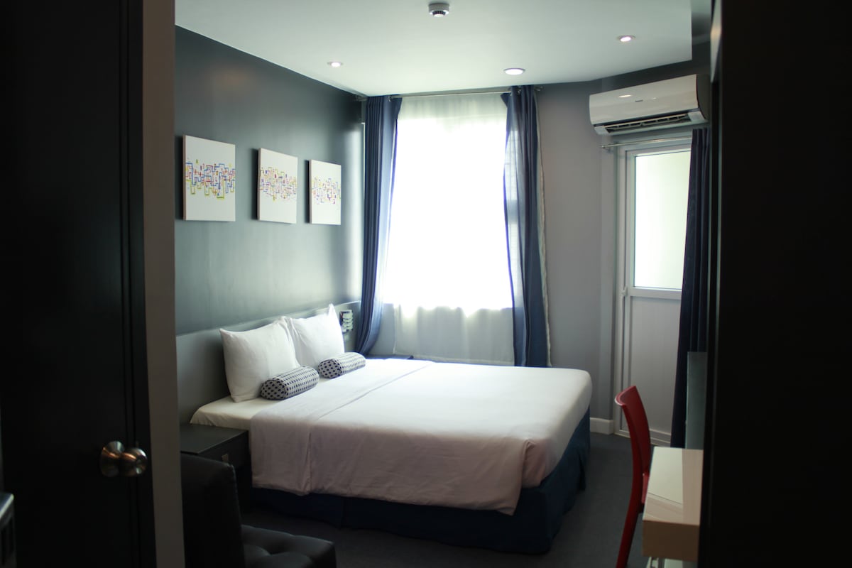 A private room is featured with a spacious bed, flanked by two cushions. Light-colored artwork decorates the dark walls. Natural light enters through the window, accented by flowing curtains. An air conditioning unit is mounted near the ceiling, and a small desk is positioned against one wall.