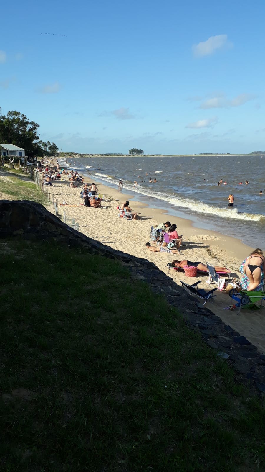 A sandy beach is lined with people enjoying the sun and water. Some are lounging on colorful towels, while others wade in the gently lapping waves. The shoreline is framed by lush greenery and a glimpse of a building can be seen in the background.
