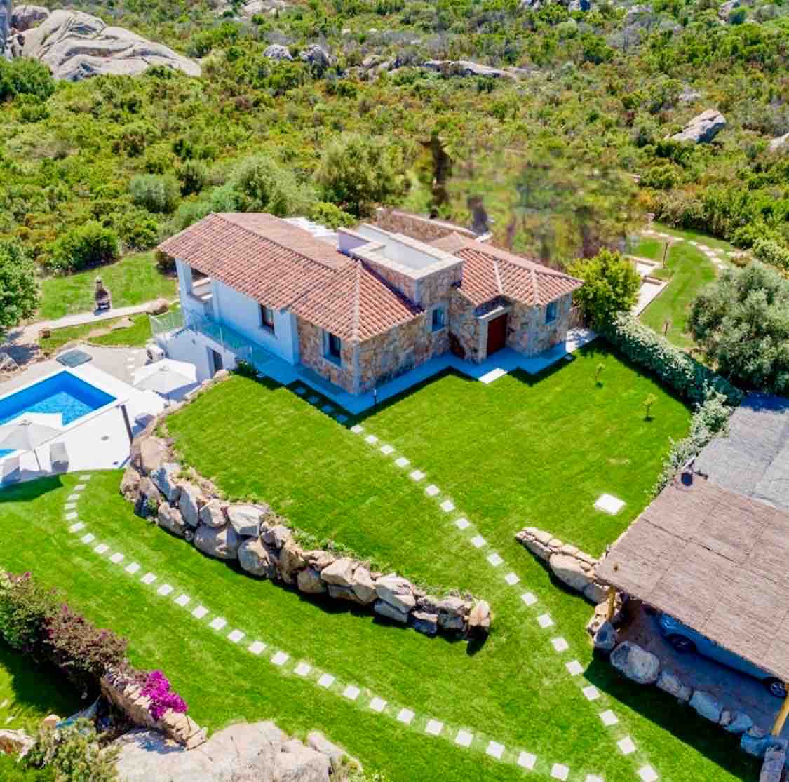 Aerial view of the villa set within a spacious green lawn, outlined with stone pathways. A private pool is visible beside the house, surrounded by lush vegetation. The villa's natural stone facade complements the surrounding landscape of rugged rocks and greenery.