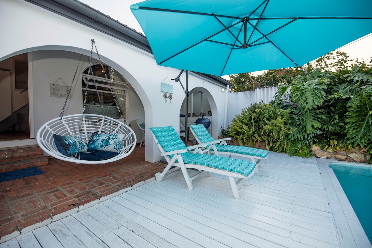 A charming outdoor space features a white wooden deck with two lounge chairs adorned with turquoise cushions. A hanging chair is positioned nearby under a large umbrella, offering shade. Lush greenery surrounds the area, adding to the tranquil atmosphere.