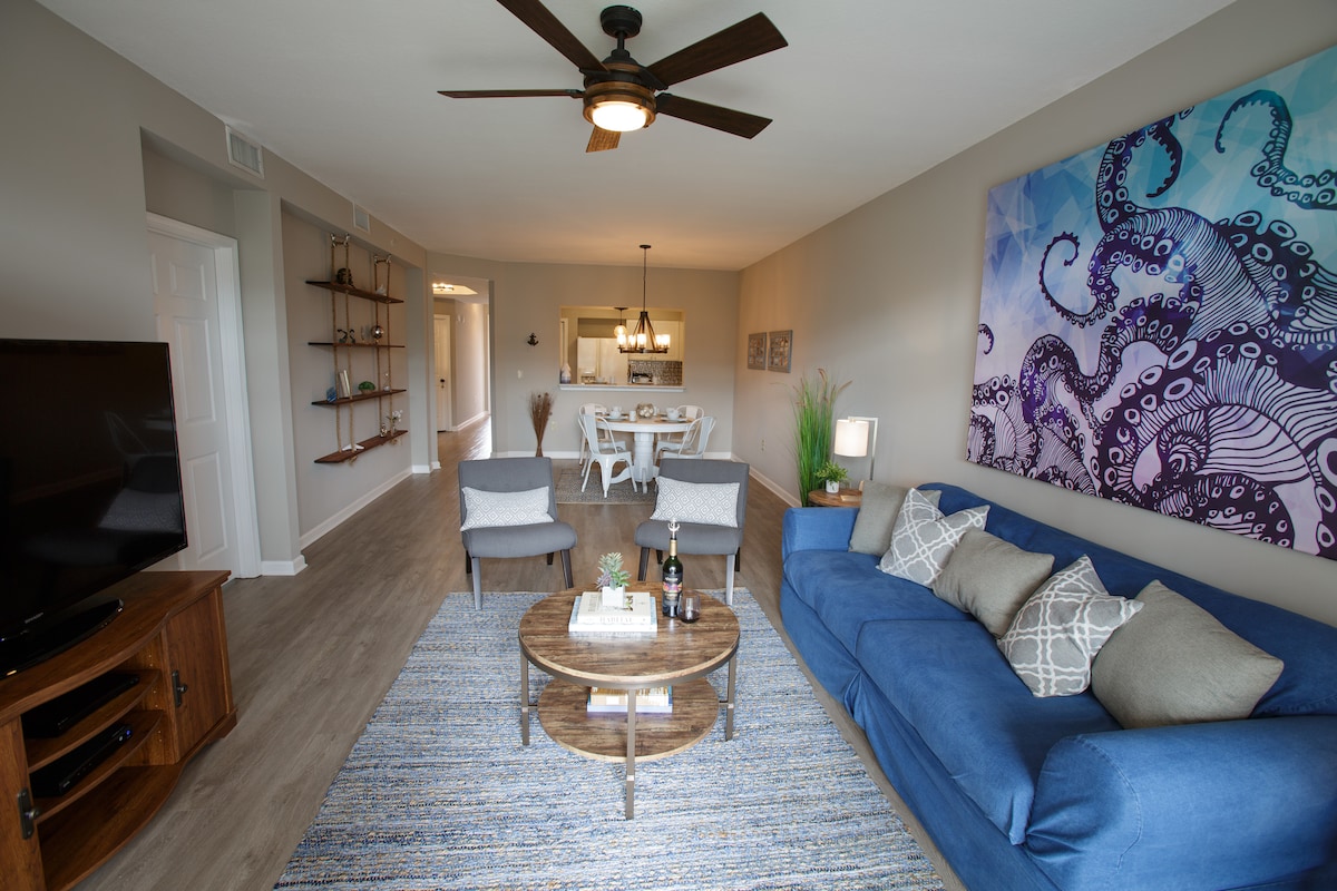 The living area features a spacious blue sofa with decorative pillows, complemented by two accent chairs and a round coffee table. A flat-screen TV is positioned against a neutral wall. An octopus-themed artwork adds a vibrant touch above a light wood console.