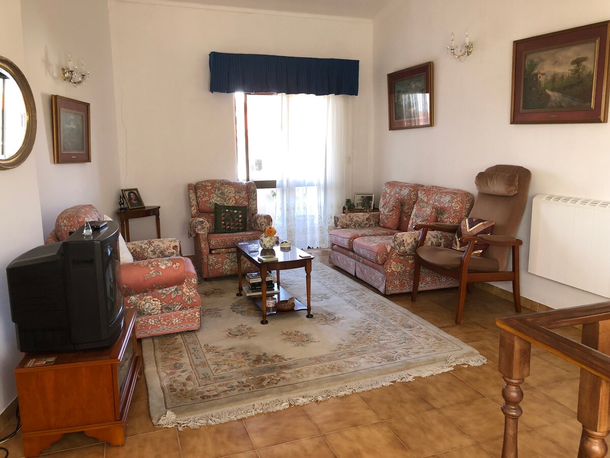 A cozy living room is presented with floral-patterned sofas and a wooden table at the center. A small television sits on a wooden stand. Framed artwork adorns the walls, while a large window allows natural light to fill the space, complemented by light-colored tiled flooring.