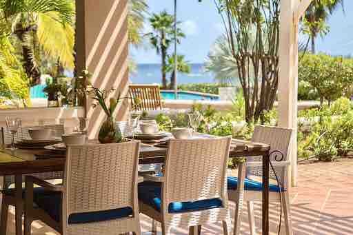 An outdoor dining area is presented, featuring a large wooden table set with dishware and seating for six. Sunlight filters through palm leaves, illuminating the space while offering views of the private pool and the sea in the background.