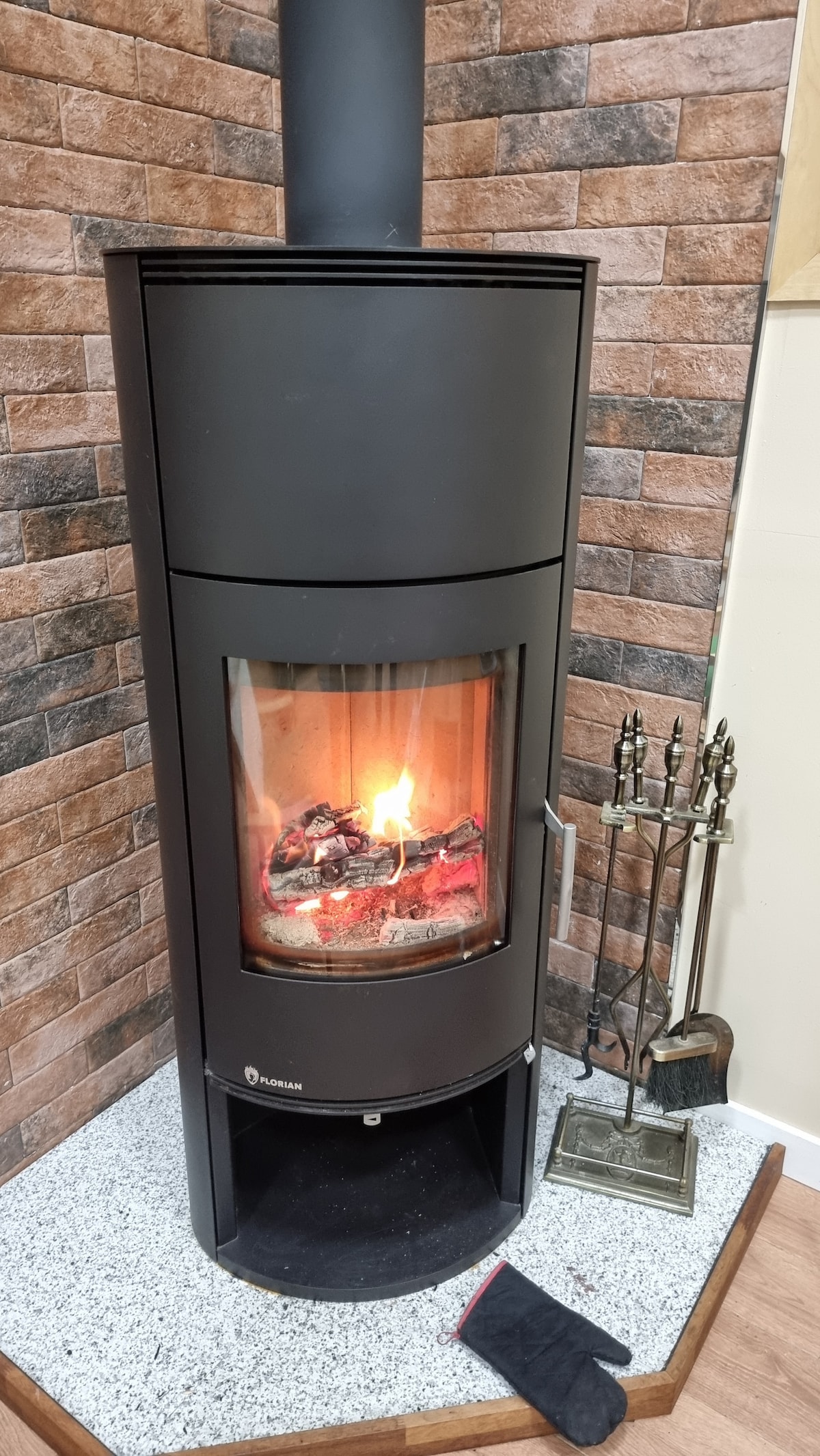 A modern wood-burning fireplace is depicted, featuring a cylindrical black design with a glass front, showcasing flickering flames and glowing embers inside. The fireplace stands on a gray stone base, complemented by a set of fire tools nearby, against a textured brown brick wall.