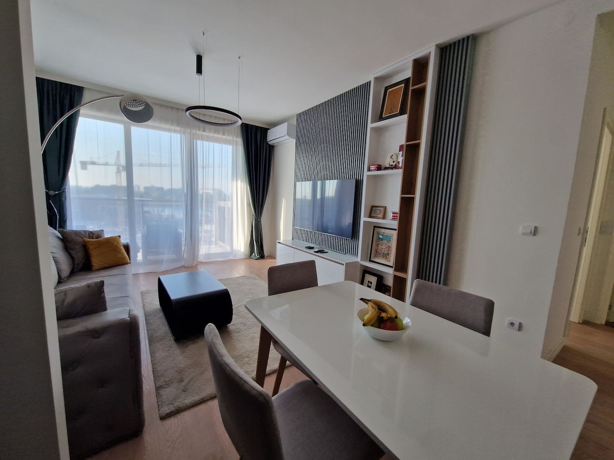 A modern and well-lit living space features a comfortable sofa, a rectangular coffee table, and a dining area with a sleek table and chairs. A large window offers views of the outside, while decorative shelves and a wall-mounted television enhance the contemporary design.