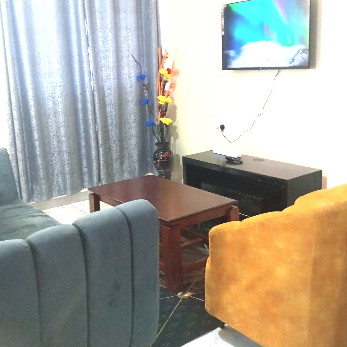 A cozy living area is presented with a dark wooden coffee table at its center, flanked by a blue sofa and an orange armchair. A flat-screen television is mounted on the wall, with decorative flowers in a vase adding a touch of color near the window, covered by grey curtains.