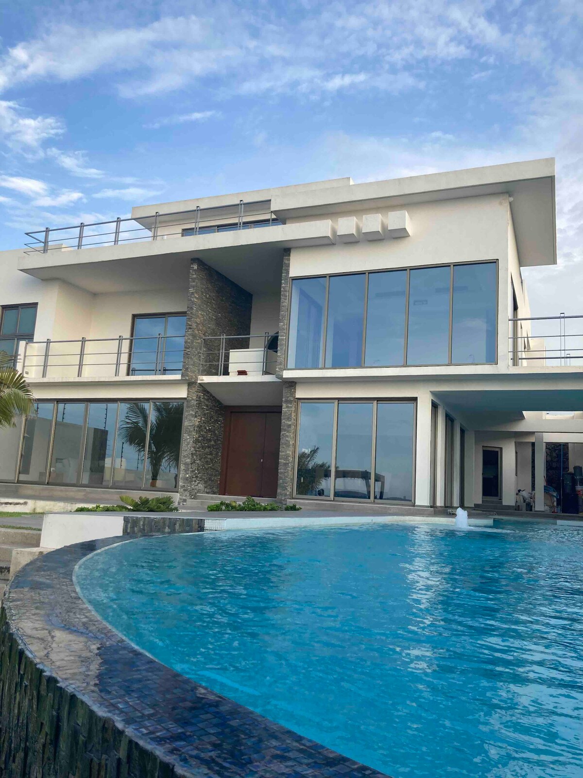 A modern villa is displayed, featuring large glass windows that offer expansive views. A pool is visible in the foreground, bordered by dark stone. The building showcases a combination of smooth and textured surfaces, with a balcony on the upper level and palm trees nearby.
