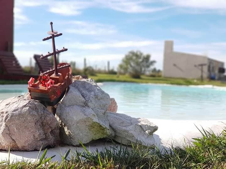 A decorative model ship is positioned atop a cluster of rocks near a natural pool. The pool, with calm water reflecting the sky, is set within a spacious outdoor area that features greenery and distant buildings.