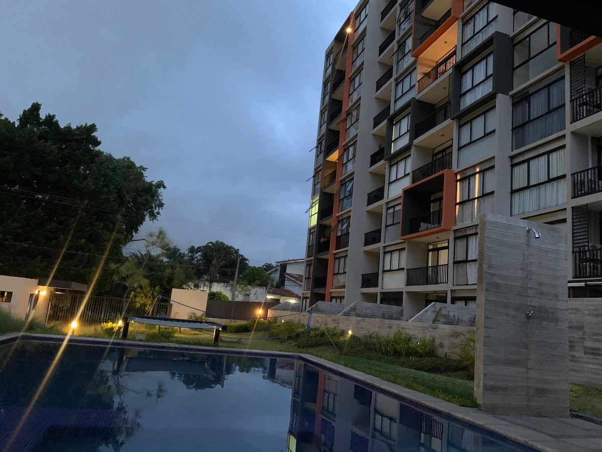 The image captures a modern condominium building illuminated softly against a twilight sky. A swimming pool reflects the surrounding greenery, while the building features a combination of large windows and balconies, enhancing the contemporary architectural design.