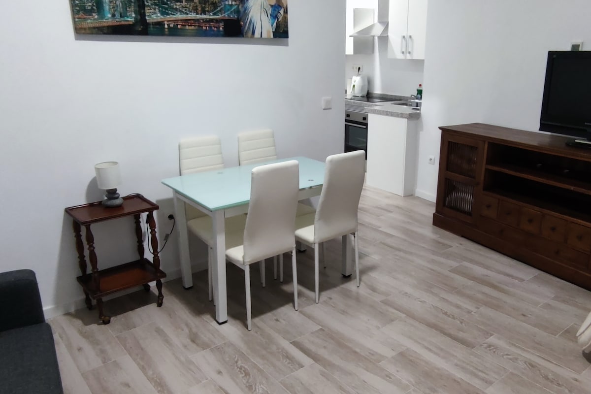 A dining area is presented with a glass-top table surrounded by four white chairs. A small wooden side table with a lamp is positioned nearby. The open kitchen is visible in the background, equipped with modern appliances, with light-colored flooring enhancing the space.