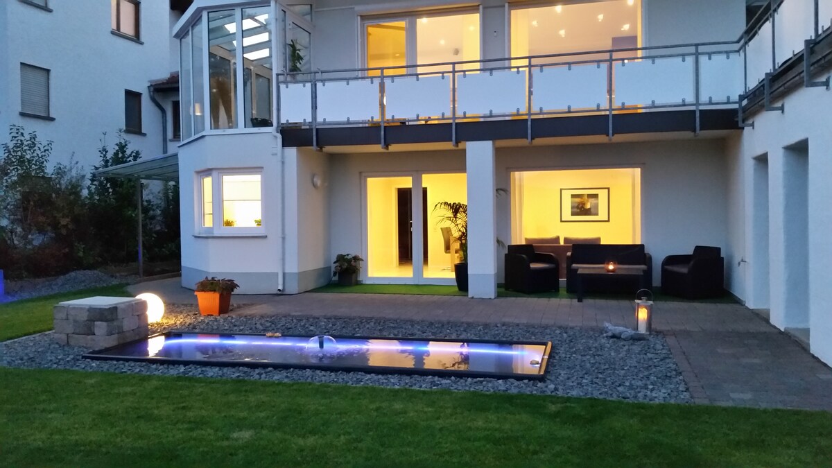 A modern outdoor space features a serene garden with a small pond illuminated by soft blue lights. Comfortable seating is arranged on a paved area, with a backdrop of large windows displaying a well-lit interior space. Lush green grass surrounds the setting.