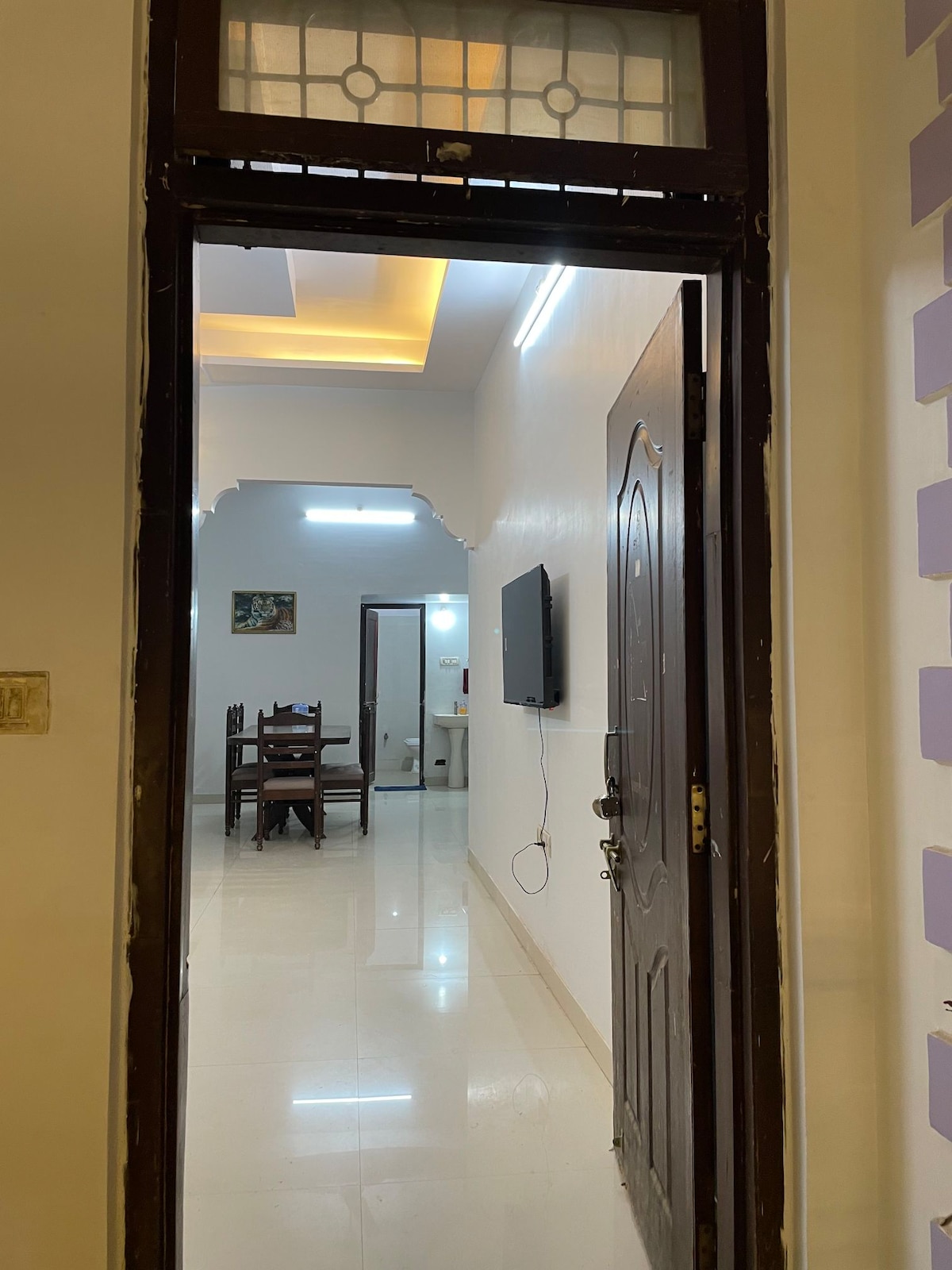 A view from the entrance reveals a well-lit drawing room featuring a dining table with four chairs. A television is mounted on the wall, and the smooth flooring reflects the natural light coming from above. The walls are designed with subtle accents.