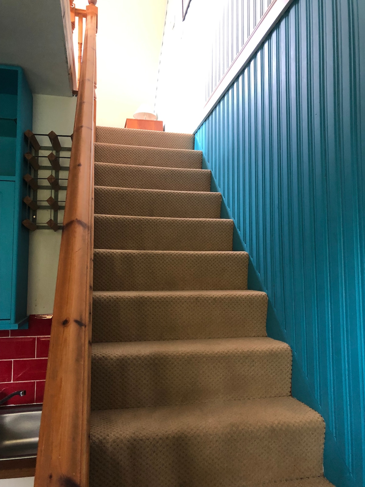 A staircase with a sturdy wooden handrail leads up to a brightly lit space. The steps are carpeted in a neutral color, and the walls are adorned with a vibrant teal paneling, creating a welcoming ascent.