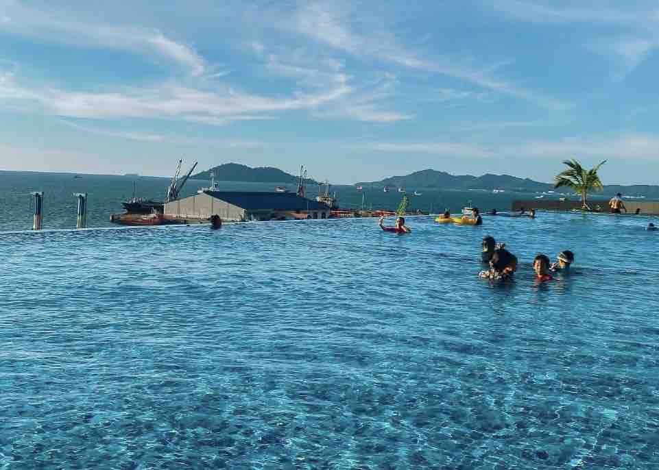 An infinity pool stretches toward the horizon, blending with the South China Sea. Several people engage in activities in the water, while boats are visible in the background. The surrounding landscape features gentle hills under a clear blue sky.