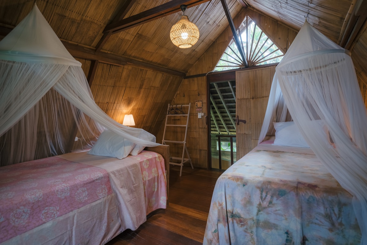 Two beds are positioned under delicate mosquito nets in a bamboo bungalow. A wooden ladder and a soft lamp are visible, providing gentle illumination. Natural light filters through the slotted window, enhancing the earthy tones of the surrounding bamboo walls.