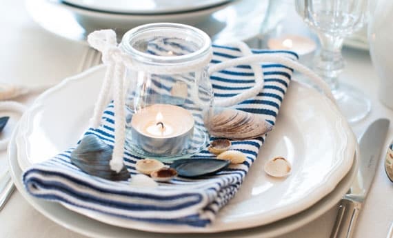 A table setting is displayed, featuring a white plate with a blue and white striped napkin underneath a small glass jar holding a candle. Decorative seashells are arranged around the plate, adding a coastal touch to the presentation.
