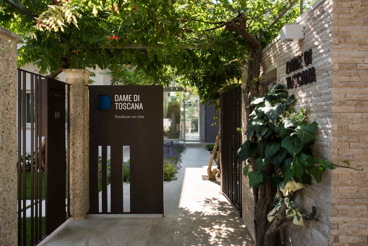 The entrance to Dame di Toscana is framed by lush greenery and decorative elements, creating an inviting pathway. The modern gate is complemented by a textured stone wall, featuring the name 'Dame di Toscana' prominently displayed. Natural light filters through the foliage above.