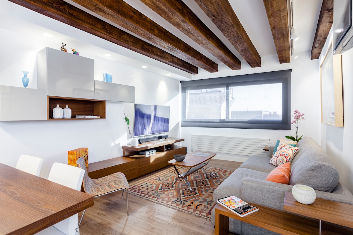 A modern living area is featured, showcasing exposed wooden beams and warm tones. A comfortable sofa is positioned beside a sleek entertainment unit with a large television. A dining table is included, complemented by stylish chairs. Decorative elements and a vibrant rug enhance the inviting space.