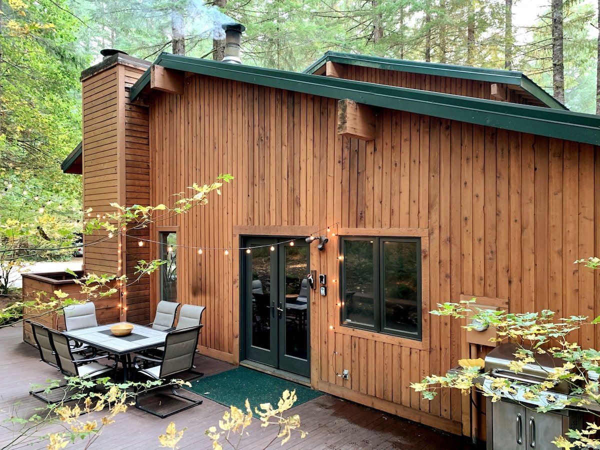 The exterior of a wooden lodge is surrounded by tall trees, showcasing natural rustic charm. A patio area features a dining table with several chairs. String lights are hung for ambiance, and a grill is positioned nearby, adding to the inviting outdoor space.