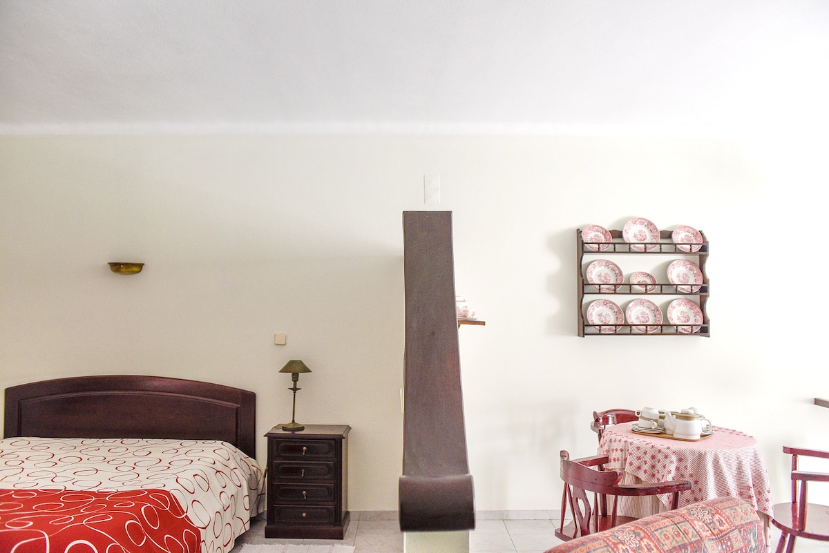 A cozy sleeping area is divided by a wooden beam, featuring a spacious bed with a circular-patterned duvet. A small bedside lamp sits on a nightstand. A dining area is located in the corner, equipped with a round table, four chairs, and decorative dishware displayed on the wall.
