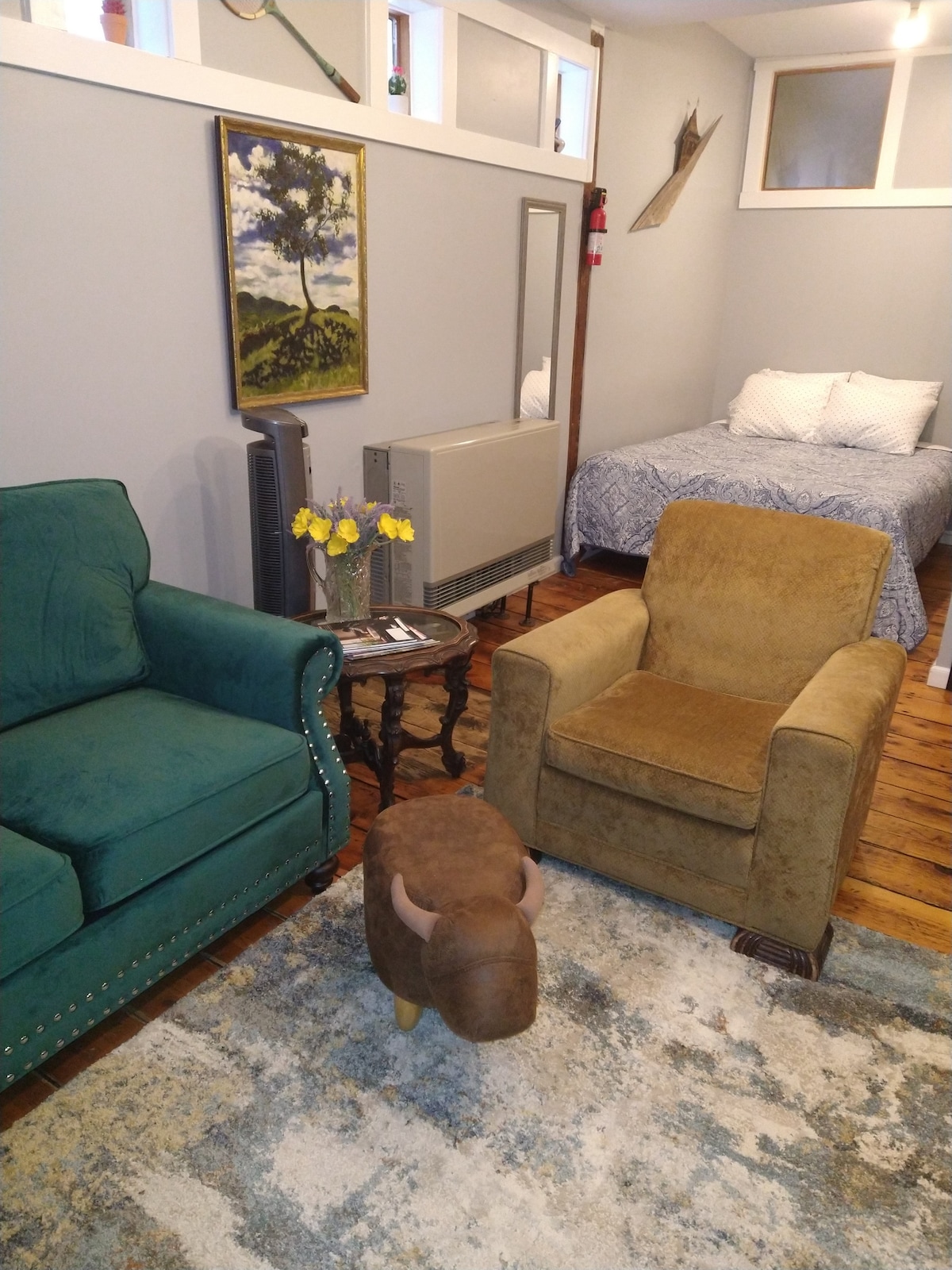 An inviting studio space is displayed, featuring a green upholstered chair, a brown armchair, and a small side table with flowers. A comfortable queen bed is positioned in the back, with light-colored bedding. A modern heating unit and a framed landscape artwork add character to the room.