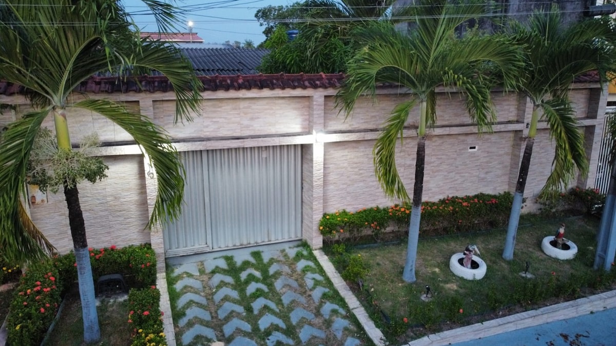 The exterior view features a well-maintained garden with neatly arranged plants and flowers. Palm trees line the pathway leading to the garage door, which is illuminated by soft lighting. A stone pathway is visible in the front yard, adding to the overall charm of the property.