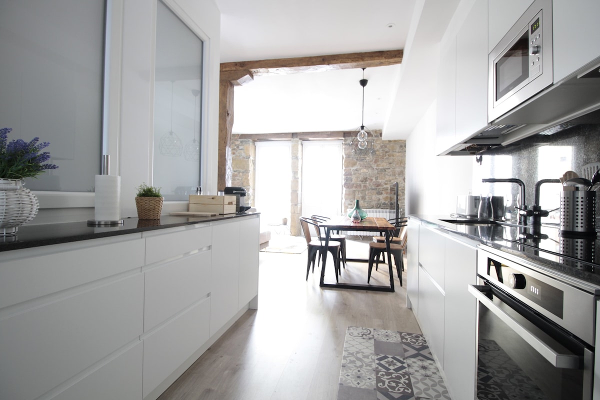 A modern kitchen features sleek white cabinetry and a dark stone countertop. Natural light fills the space through large windows, highlighting a wooden dining table with black chairs. The kitchen appliances, including a microwave and oven, are seamlessly integrated into the design.