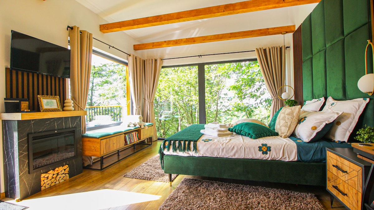 A cozy bedroom features a spacious bed with decorative pillows and throws. Large windows provide views of the surrounding greenery. A stylish fireplace and a small seating area are present, with wooden beams enhancing the room's contemporary charm.