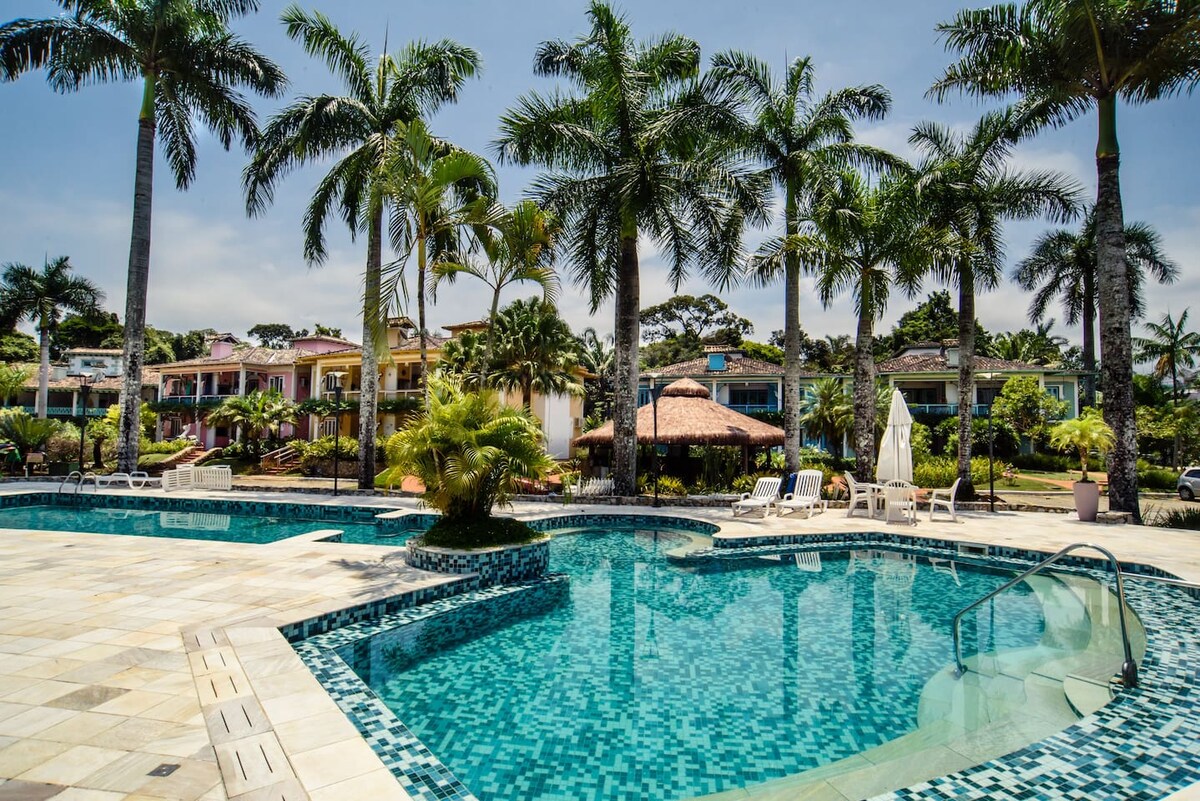 A landscaped pool area is surrounded by tall palm trees and tropical plants. Two swimming pools are visible, one featuring a curved shape. Relaxing lounge chairs are positioned nearby, inviting leisure time. A thatched-roof gazebo can be seen in the background, enhancing the outdoor ambiance.
