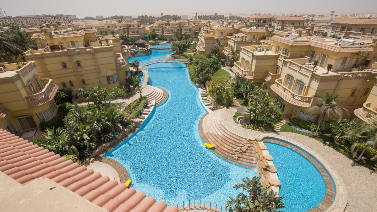 A bird's-eye view captures a winding pool surrounded by lush greenery and well-manicured landscaping. Multiple lounge chairs are positioned along the water's edge. The architecture of nearby buildings offers a harmonious blend with the natural elements of the tranquil environment.