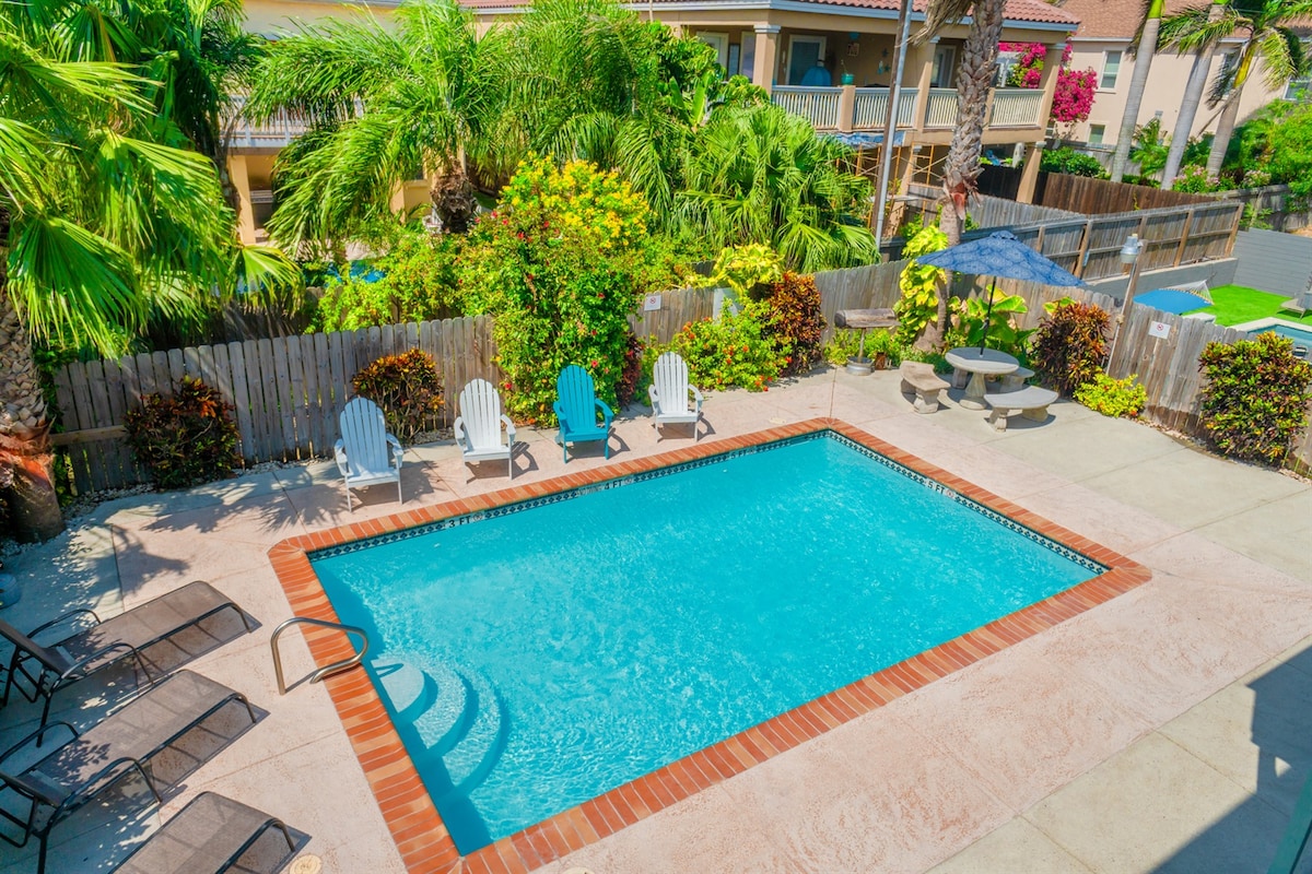 An inviting swimming pool is surrounded by lush greenery and tropical plants. Several white Adirondack chairs are positioned beside the pool, offering space for relaxation. A shaded seating area with a round concrete table and benches can be seen to the side, providing an area for outdoor dining.