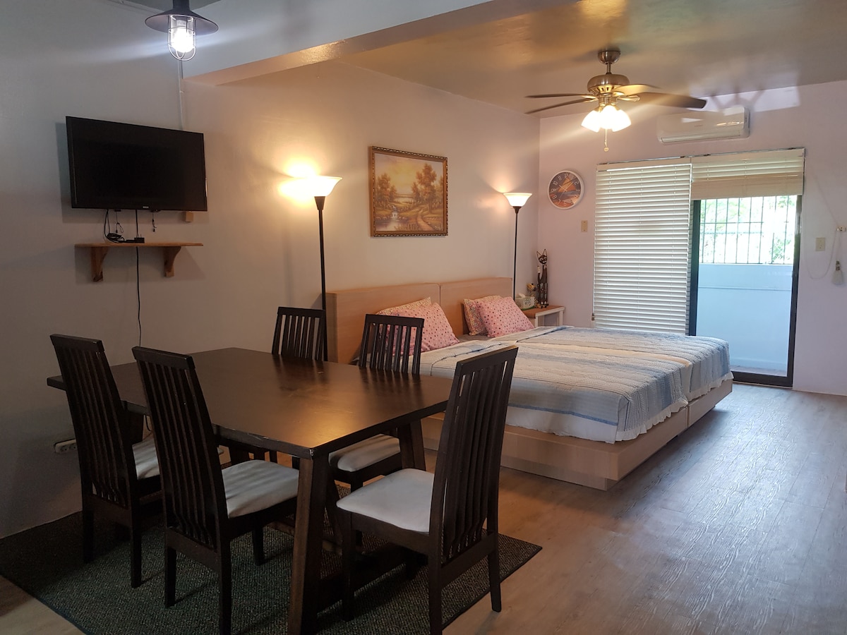 A spacious sleeping area features a light-colored bed with patterned pillows and overhead ceiling fans. A dining table with dark wooden chairs occupies one side, while a mounted television hangs on the wall. Natural light enters through a sliding door leading to an outdoor area.