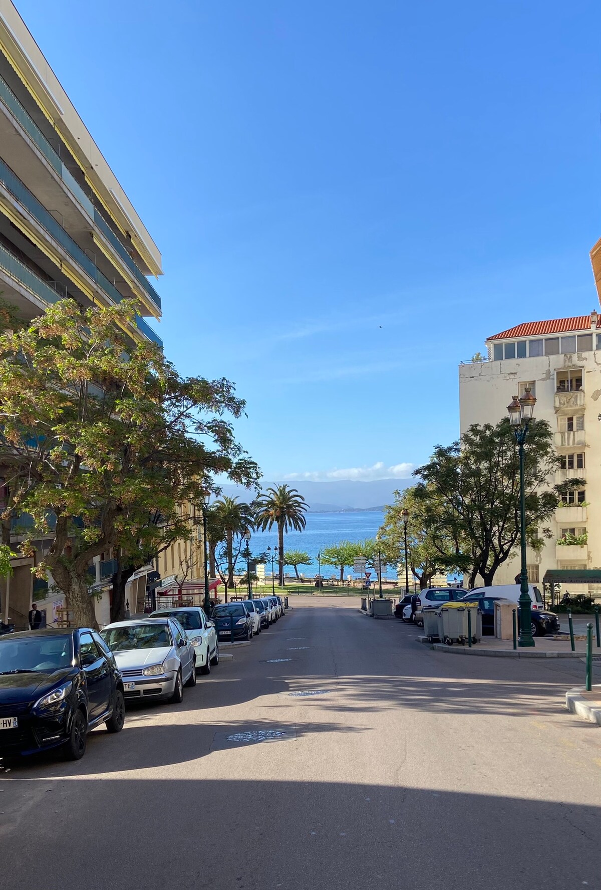 A broad avenue lined with parked cars leads to a view of the coastline. Palm trees dot the landscape, and the azure sea is visible in the distance under a clear blue sky, creating a serene coastal atmosphere.