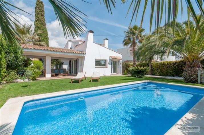 A private pool glimmers in the sun, surrounded by a well-maintained garden featuring palm trees and lush greenery. A covered outdoor seating area provides shade, while two lounge chairs are positioned near the water. The house's white exterior contrasts with the vibrant blue sky.