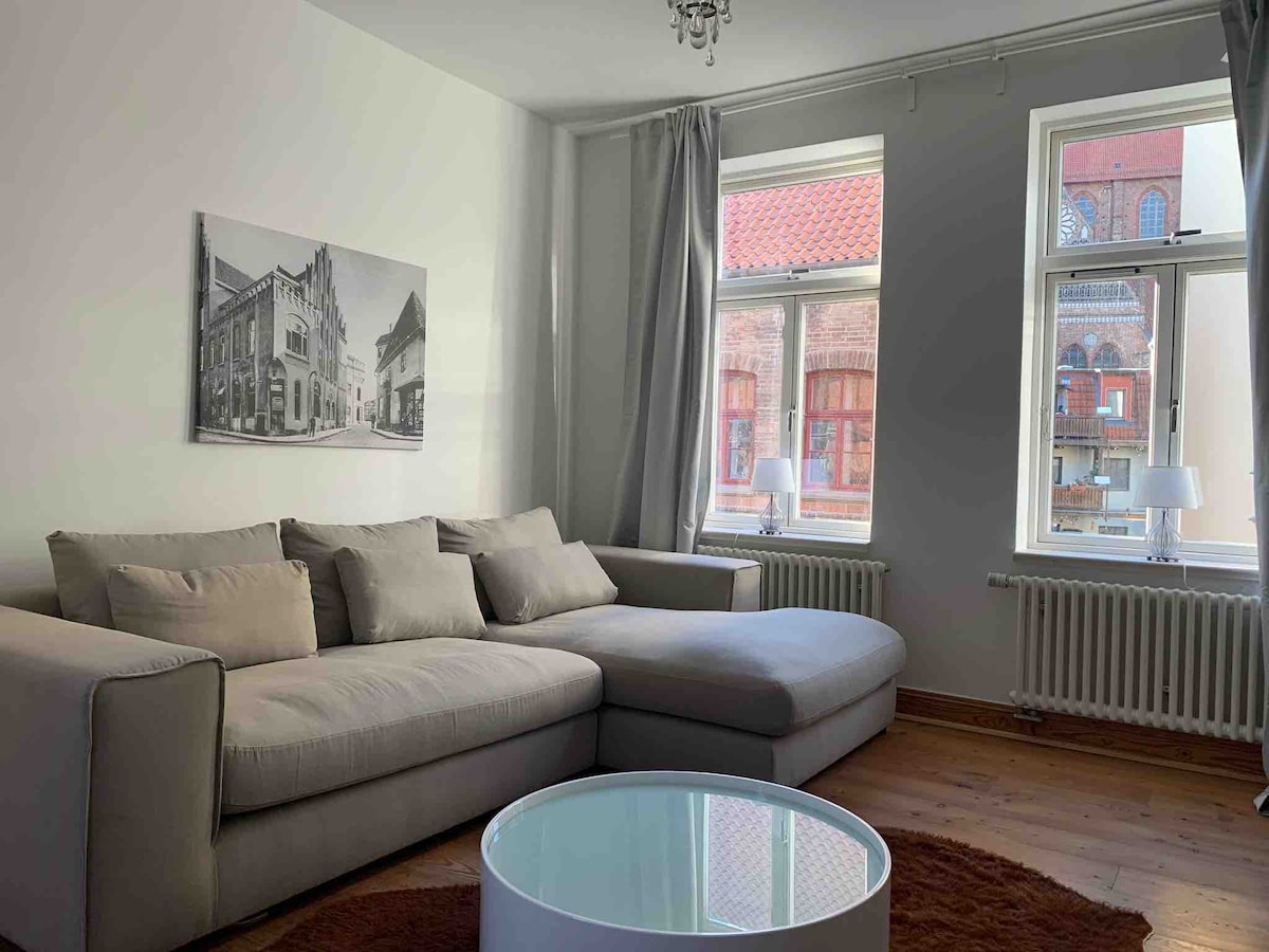 A spacious living area features a light-colored sectional sofa and a round coffee table on a soft rug. Two windows allow natural light to fill the room, with decorative curtains framing the views of the historic buildings outside.