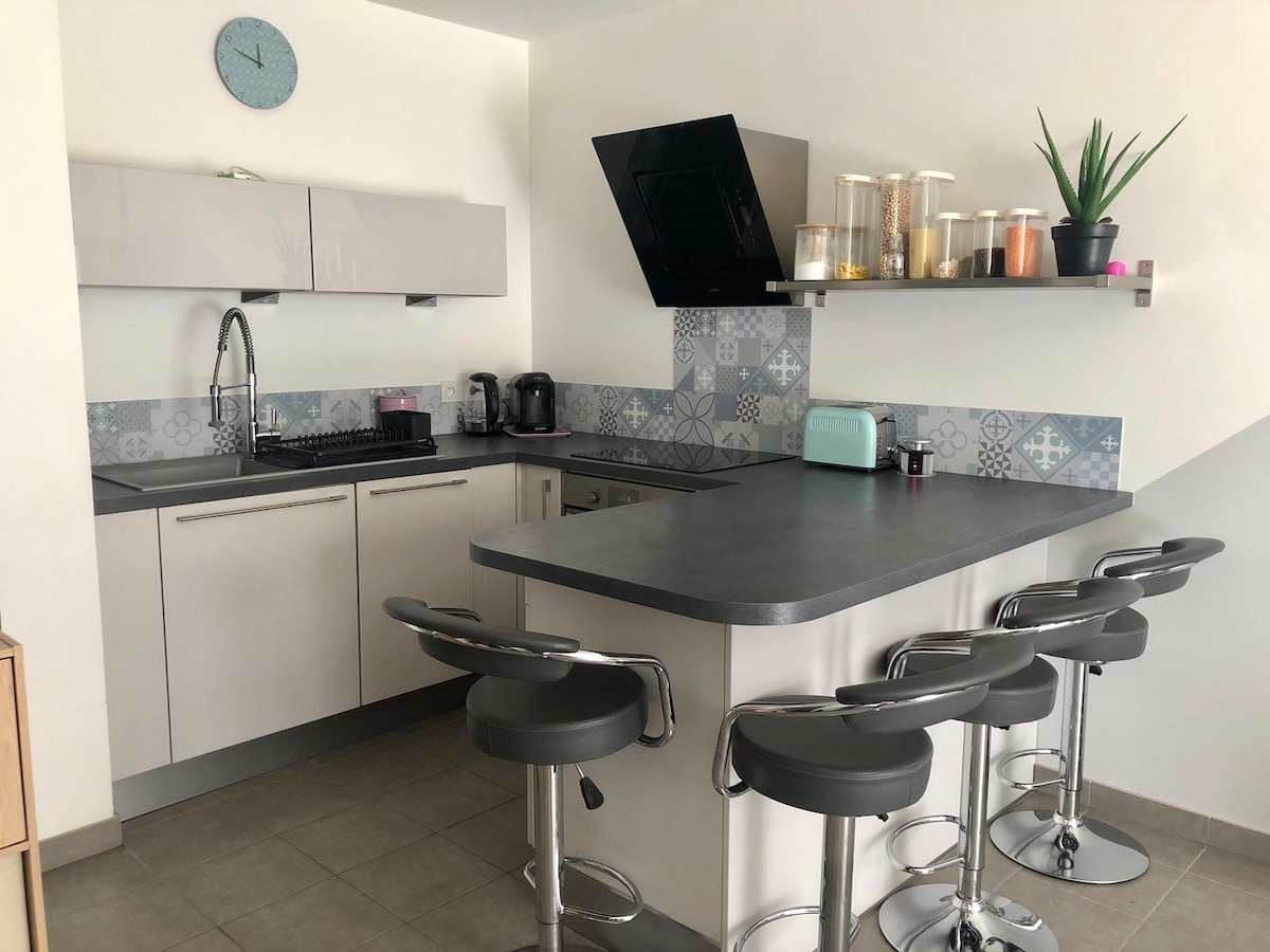 The compact kitchen features modern cabinetry with a gray countertop. A breakfast bar with stools invites casual dining. Wall shelving displays decorative plants and jars, while a blue and white patterned backsplash adds visual interest. Essential appliances are neatly arranged along the countertop.