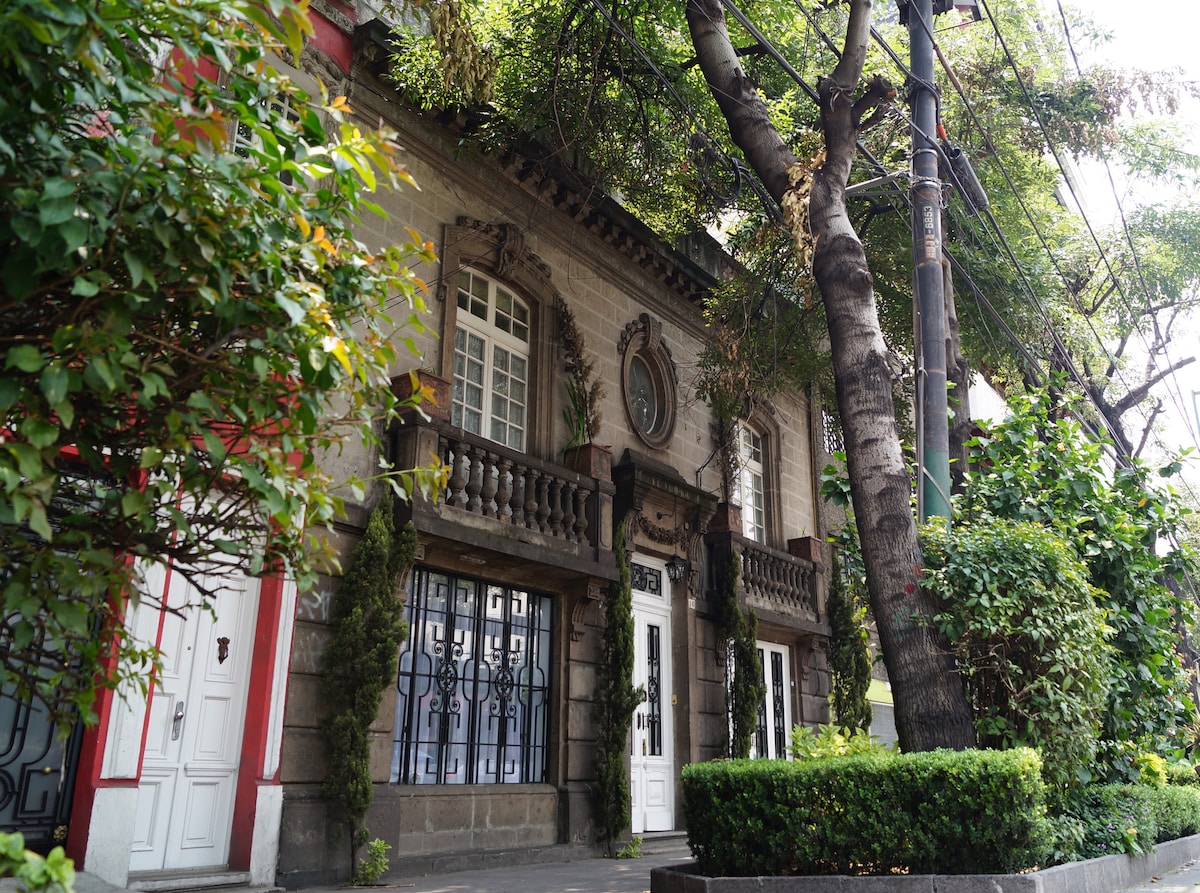 The exterior of a historic 1920s Art Nouveau mansion is displayed, featuring grand balconies and decorative stonework. Lush greenery surrounds the building, enhancing the inviting atmosphere. A tree stands prominently near the entrance, with well-maintained hedges lining the walkway.