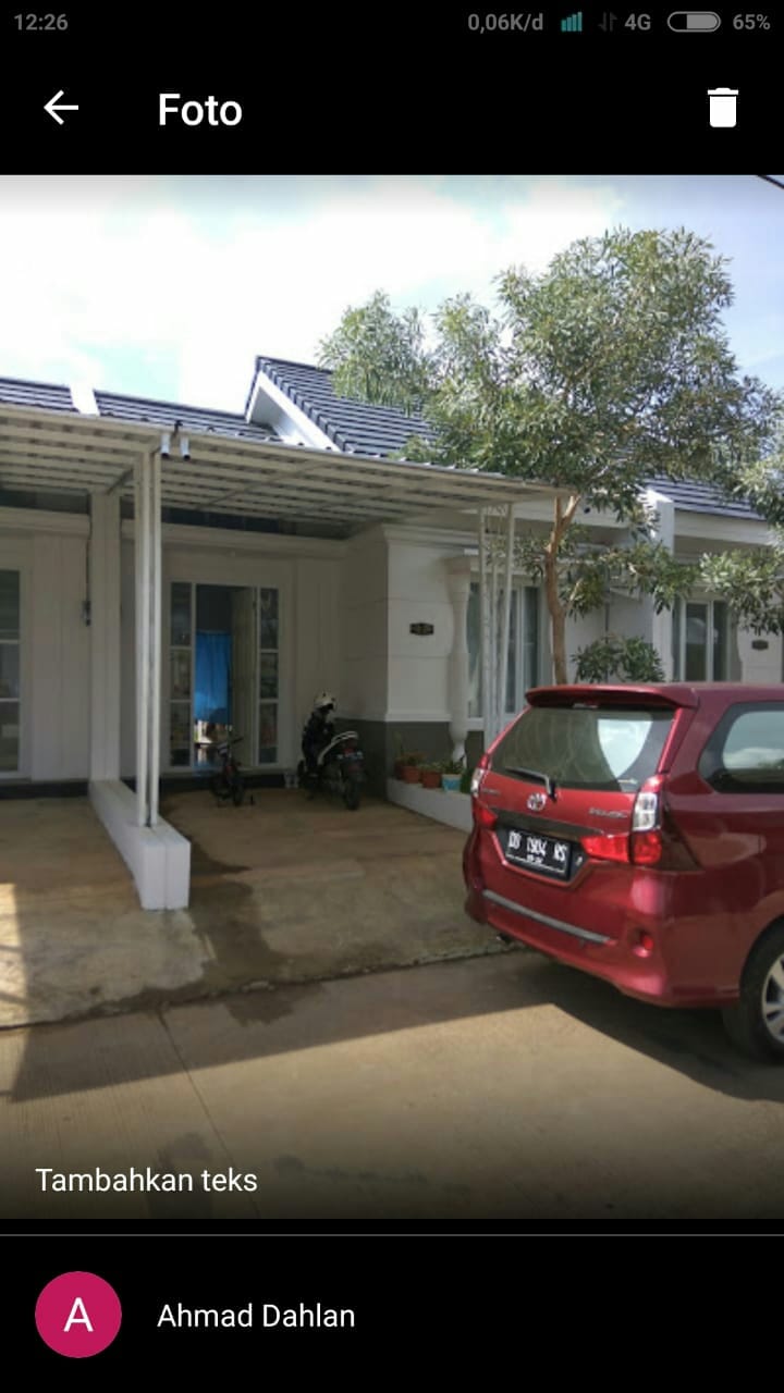 The exterior of a comfortable residential home is shown, featuring a covered parking area for a small car. A motorcycle is parked nearby. Lush greenery is visible alongside the façade, with large windows allowing natural light into the house.