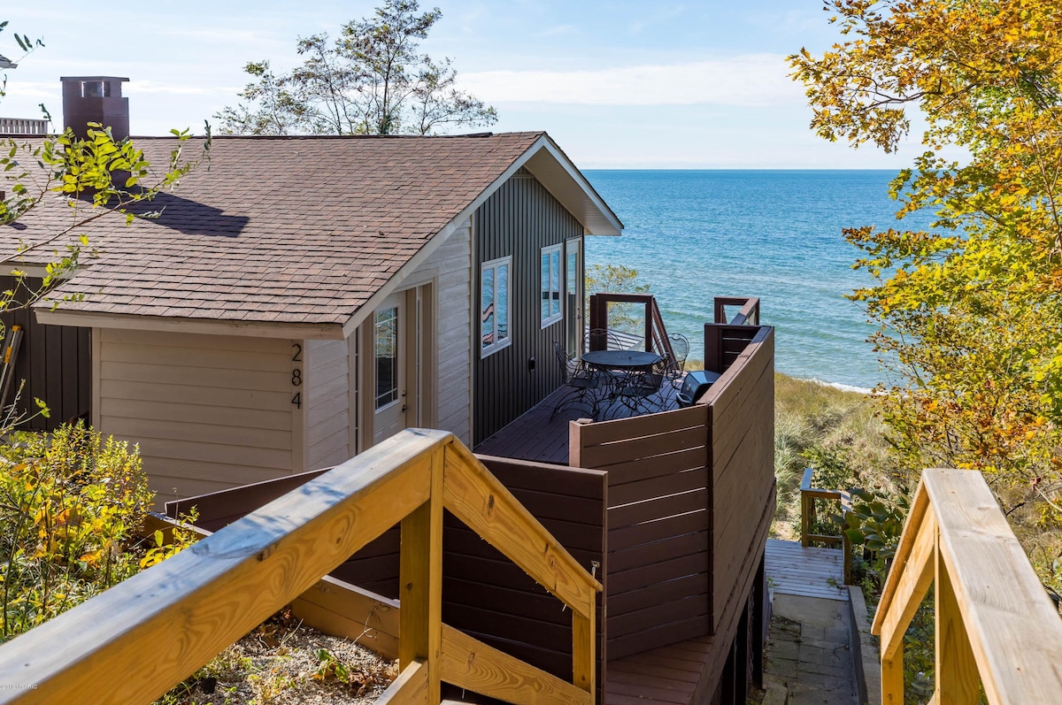 The exterior of the remodeled beach retreat is shown, featuring a simple brown and cream facade. A wooden staircase leads down through lush vegetation to a raised deck, which offers views of Lake Michigan and a seating area that overlooks the shoreline.