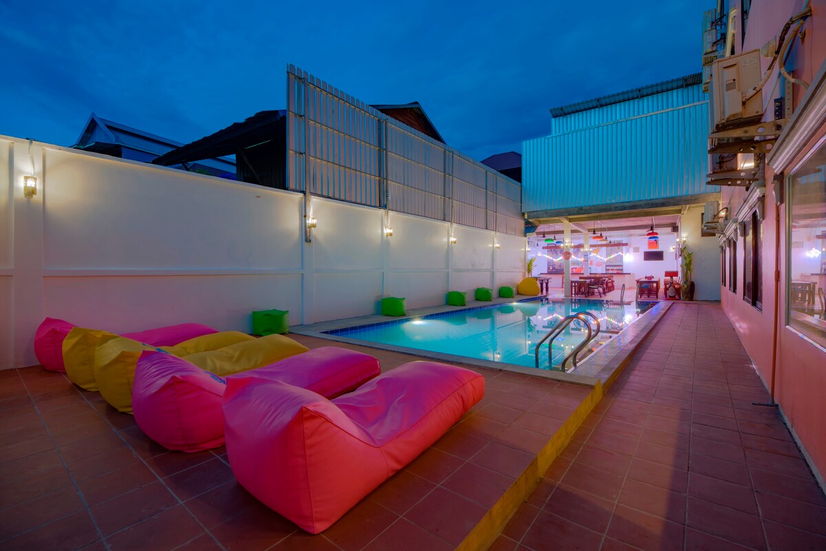 A vibrant outdoor area features a rectangular swimming pool surrounded by colorful bean bags in pink and green. Soft lighting from the surrounding structures creates a relaxed evening ambiance, with additional seating visible near the poolside.