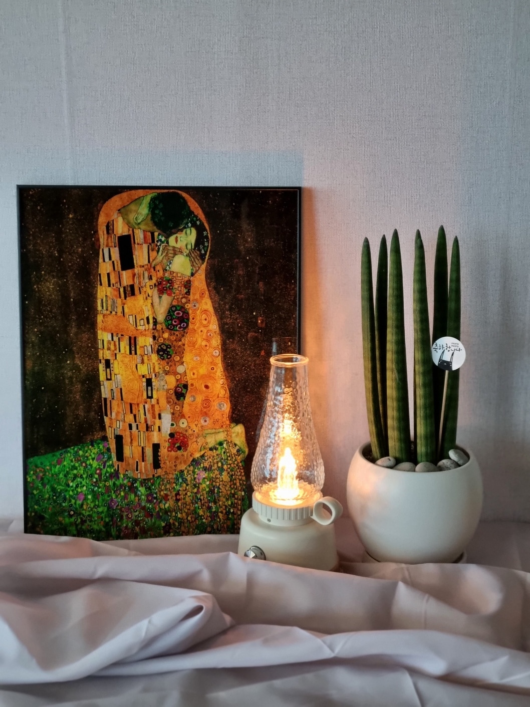 A warm image features a vibrant artwork on the left, depicting an embracing couple surrounded by ornamental details. To the right, a potted plant with long green leaves stands beside a lamp emitting a gentle glow, creating a serene atmosphere on a softly draped surface.