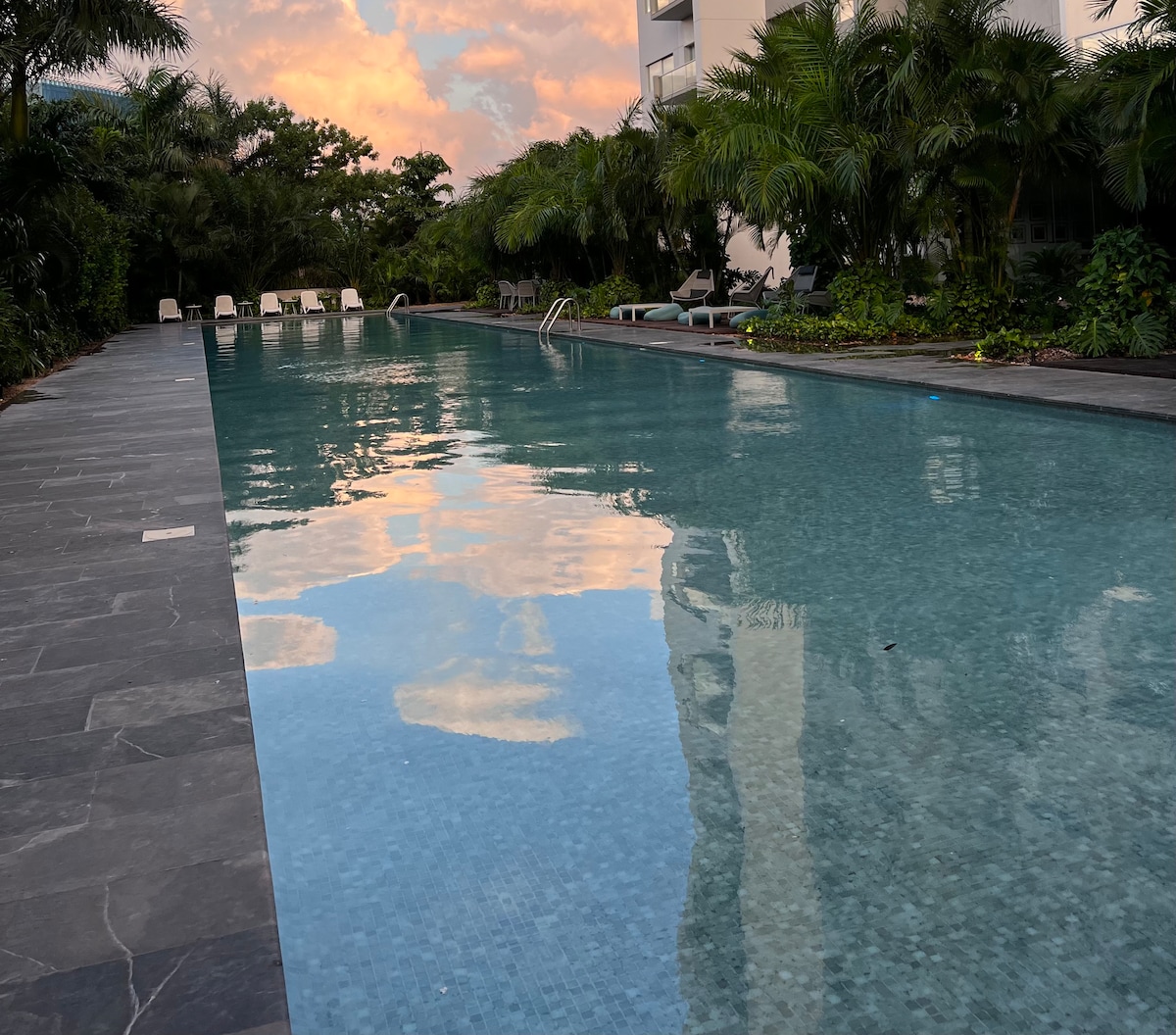 A tranquil pool is surrounded by lush greenery and palm trees. The clear water reflects the soft colors of the sky during sunset, creating a serene atmosphere. Lounge chairs are positioned along the edge, inviting relaxation and enjoyment.