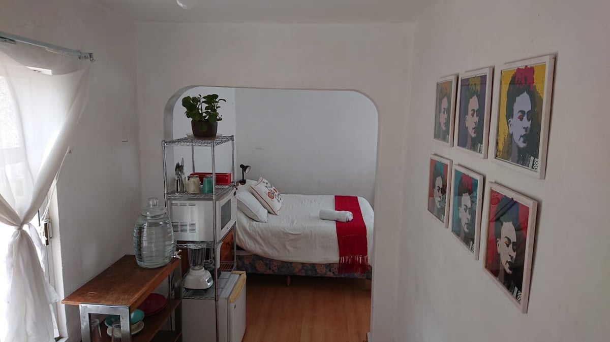 The room features a spacious bed with a white coverlet, framed by light walls. A small kitchen section is visible with a shelf holding kitchenware and a clear water container. Colorful wall art showcasing portraits adds character, while natural light enters through a nearby window.