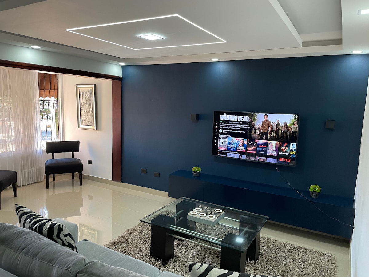 A modern living area features a dark blue accent wall with a large television displaying streaming options. A glass coffee table is centered on a light rug, surrounded by a comfortable sofa and two chairs. Natural light filters through sheer curtains.