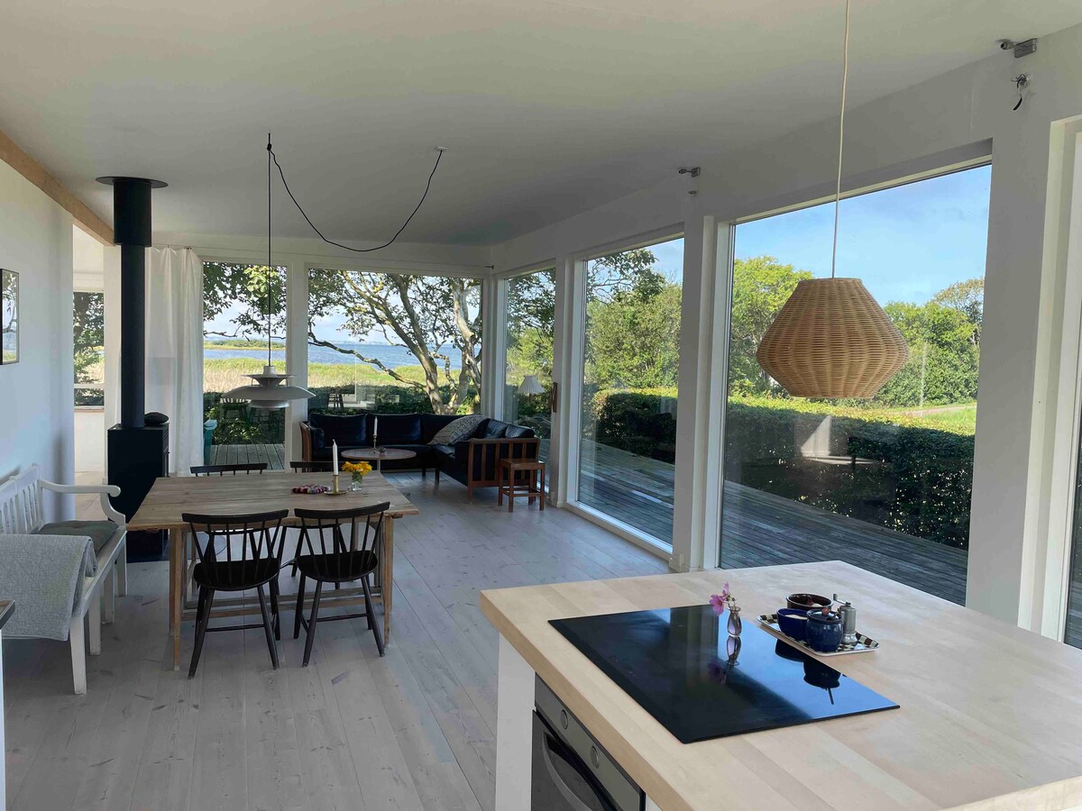 An open kitchen and dining area are highlighted by large windows offering a view of the outdoors. A wooden dining table is accompanied by black chairs, while a light fixture hangs overhead. The kitchen features a smooth countertop with cooking essentials.