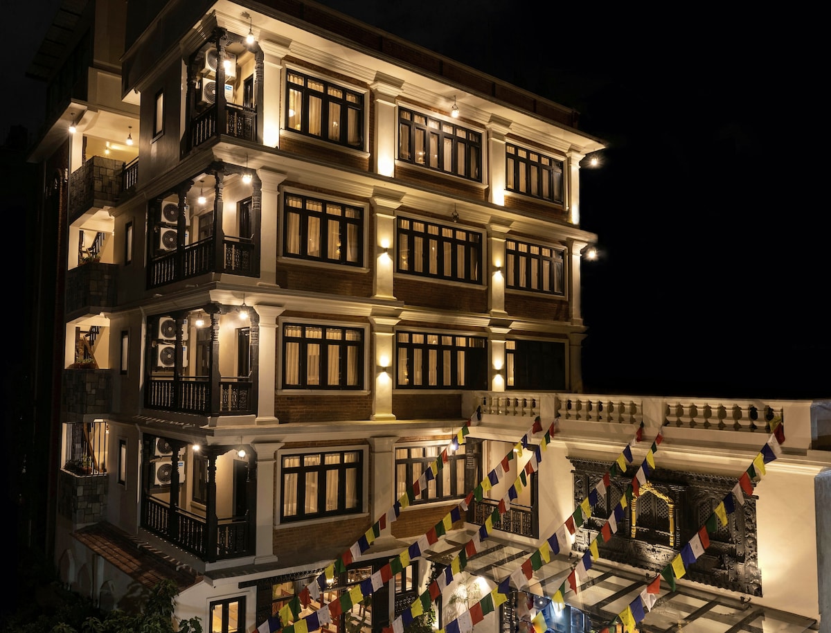The heritage building showcases a four-story structure illuminated against the night sky. Decorative balconies adorn the facade, framed by glowing windows. Colorful prayer flags are displayed in the foreground, leading to a welcoming entrance.