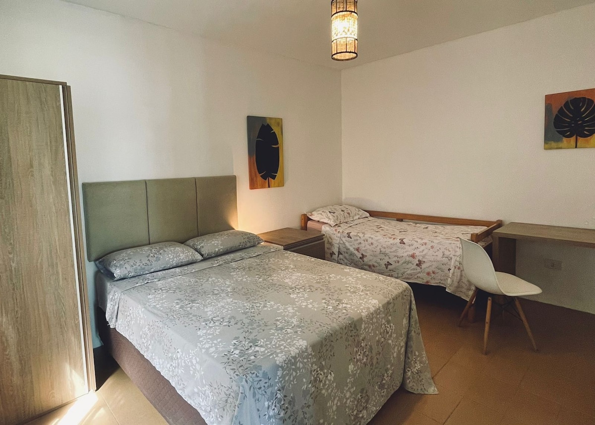 The sleeping area features a spacious bed with a light floral patterned coverlet, alongside a single bed with a simple design. Two pieces of artwork adorn the wall, and a small desk with a chair is positioned nearby.