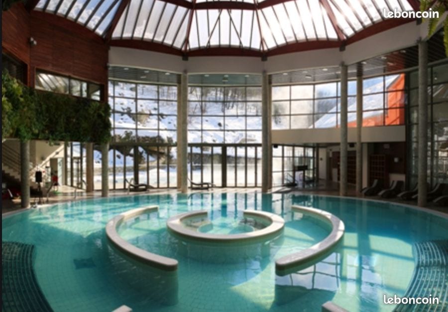 An indoor swimming pool features a circular layout with seating areas around its perimeter. Large windows allow natural light to illuminate the space, providing views of the surrounding snowy landscape. Lush greenery decorates the walls, enhancing the serene atmosphere.