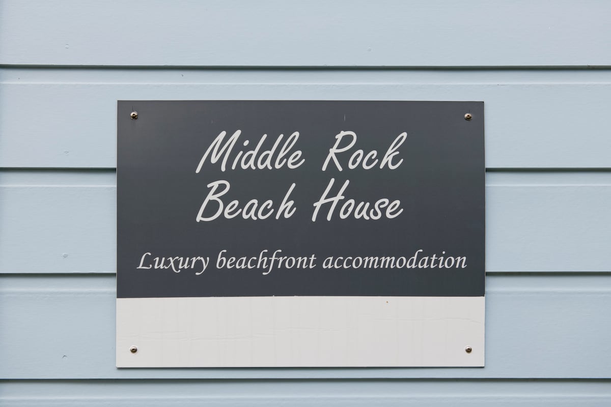 A sign is mounted on a light blue exterior wall, displaying the name 'Middle Rock Beach House' in elegant white lettering. Below, the phrase 'Luxury beachfront accommodation' is presented in a smaller font, emphasizing the inviting nature of the property.
